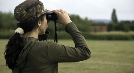 Woman with a pony tail looking away through binoculars wearing a camouflage print cap