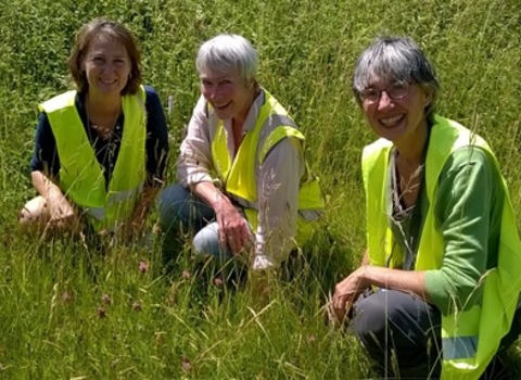Three women wearing hi-vis waistcoats kneeling in long grass smiling at camera