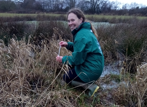 Woman in green coat crouching in long vegetation, turning to smile at camera