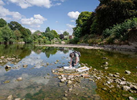 View along wide, low river in summer with man in centre kneeling over a white tray