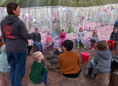 Group of children sitting around a fire on tree stumps under a canopy 