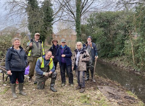 Group of seven people in outdoor clothing stood beside a small stream; trees and hedge behind; winter