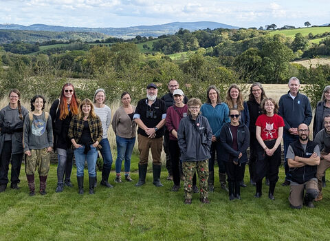 Around 25 people stood on a lawn for a group photo with rural landscape in background